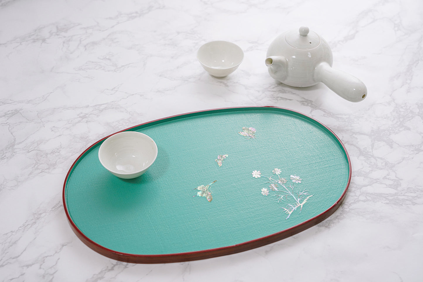 A teal oval tray with mother-of-pearl floral design, alongside a white teapot and two cups on a marble background.