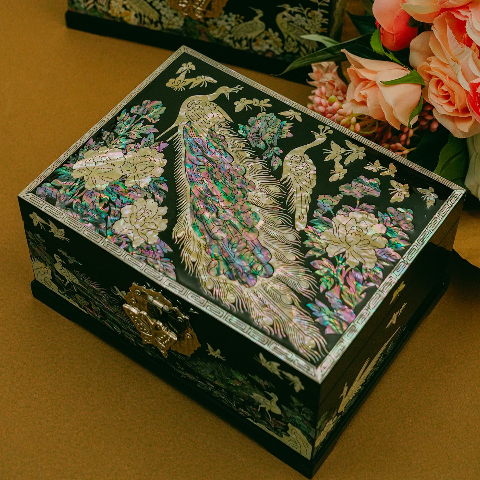 A Mother of Pearl jewelry box with peacock design is placed on a table beside pink flowers, its vibrant inlays contrasting with the warm background.
