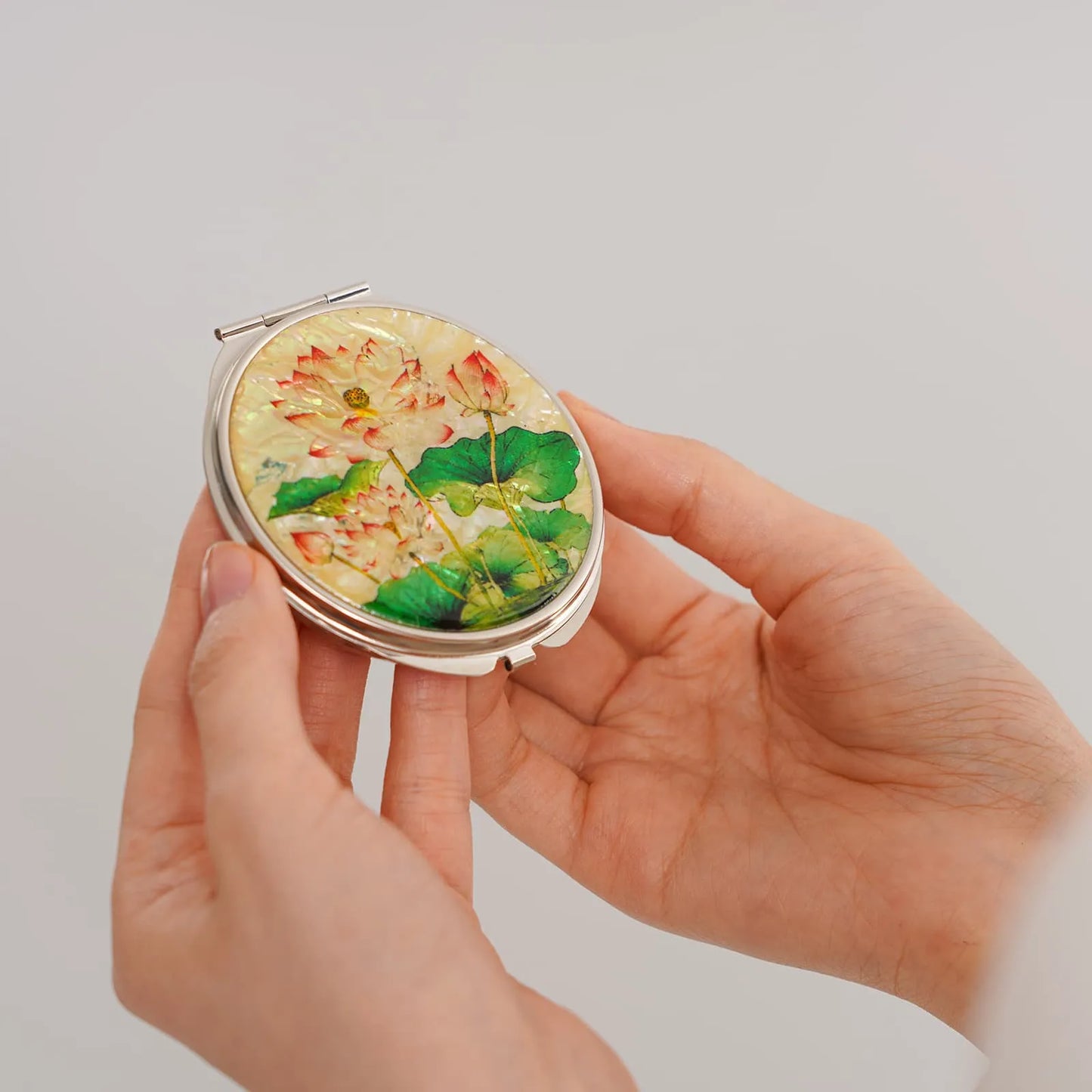 A compact mirror featuring a decorative lid with detailed lotus flowers and leaves. The flowers are primarily white with pink accents, while the leaves