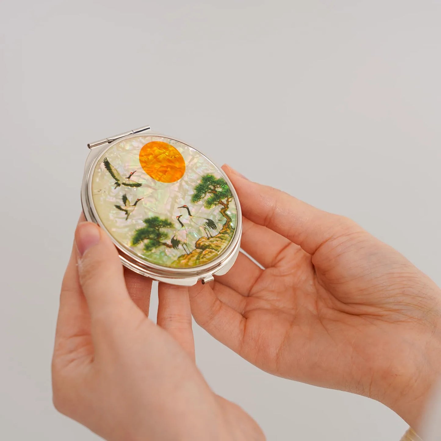 A compact mirror featuring a traditional East Asian design, with cranes, trees, and a glowing orange sun