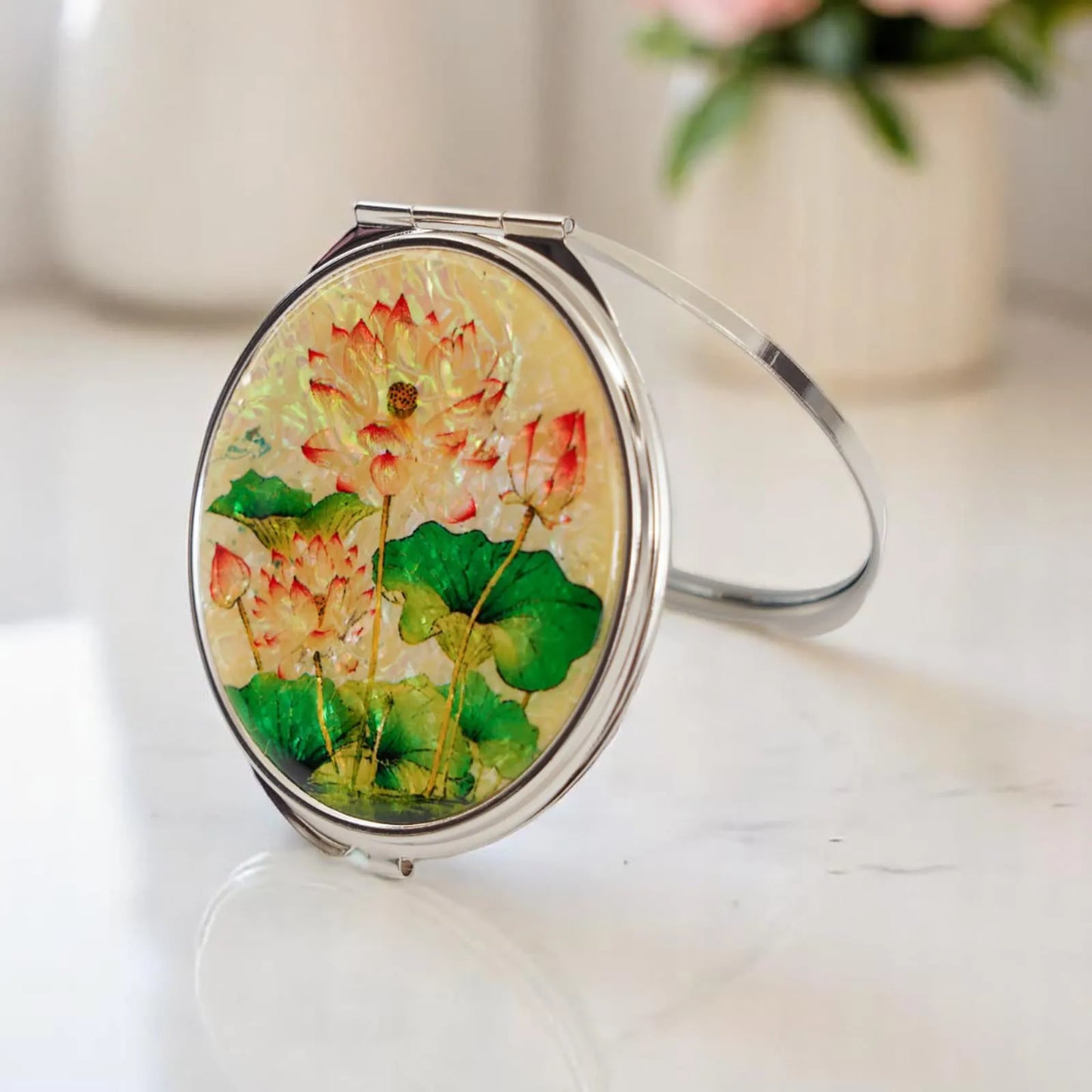 A compact mirror featuring pink lotus flowers and green leaves, set against a soft pearlescent background