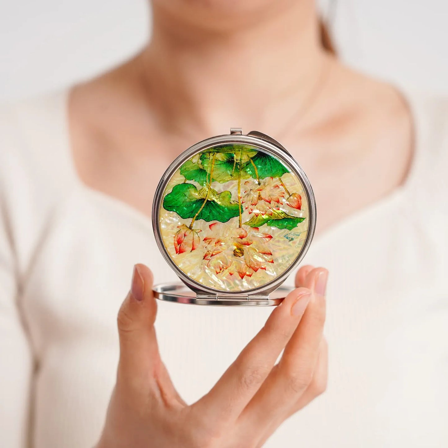 A compact mirror with a floral design featuring green leaves and light-colored flowers, held by a person in a white top