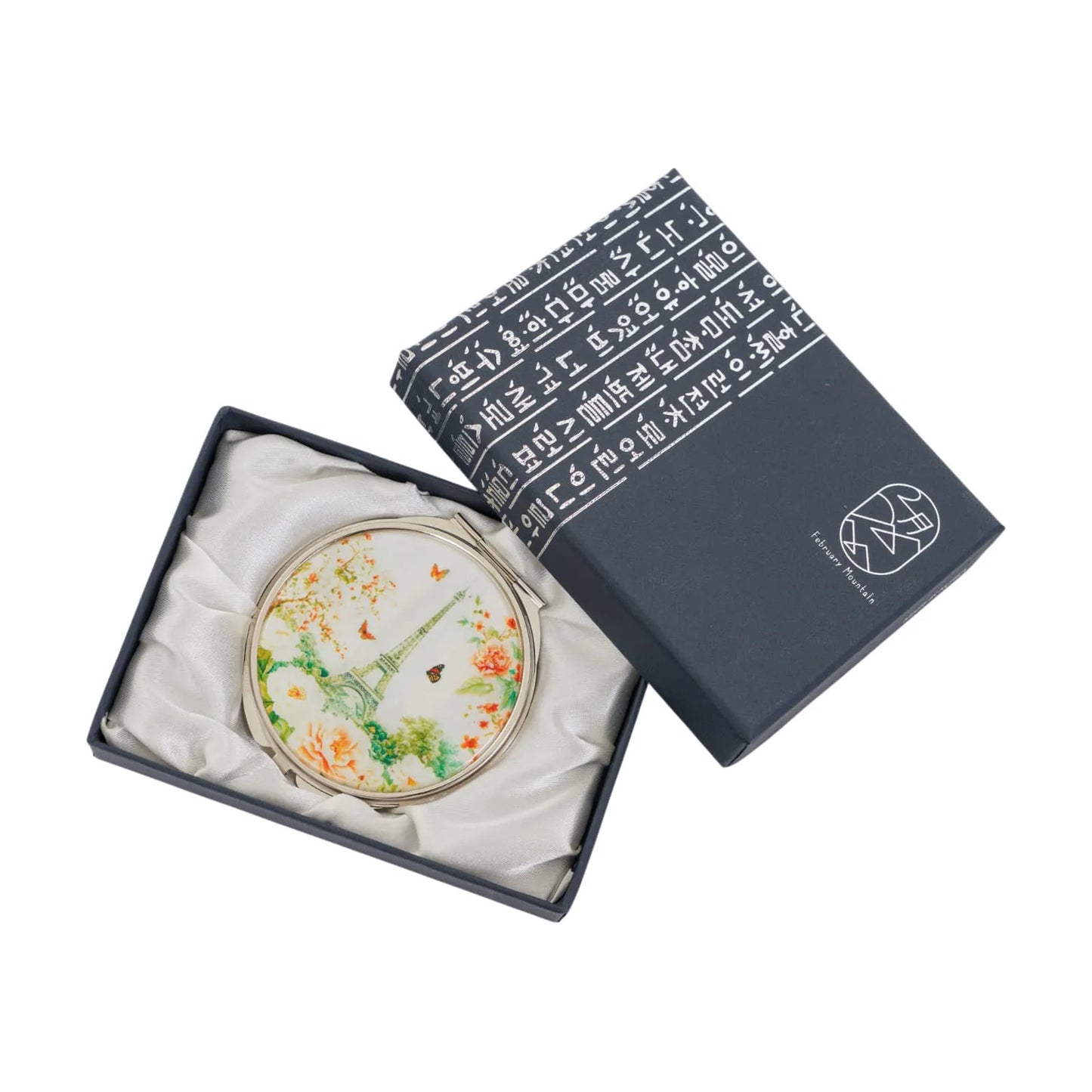 A decorative compact mirror with an Eiffel Tower design in a blue box with white text and logo