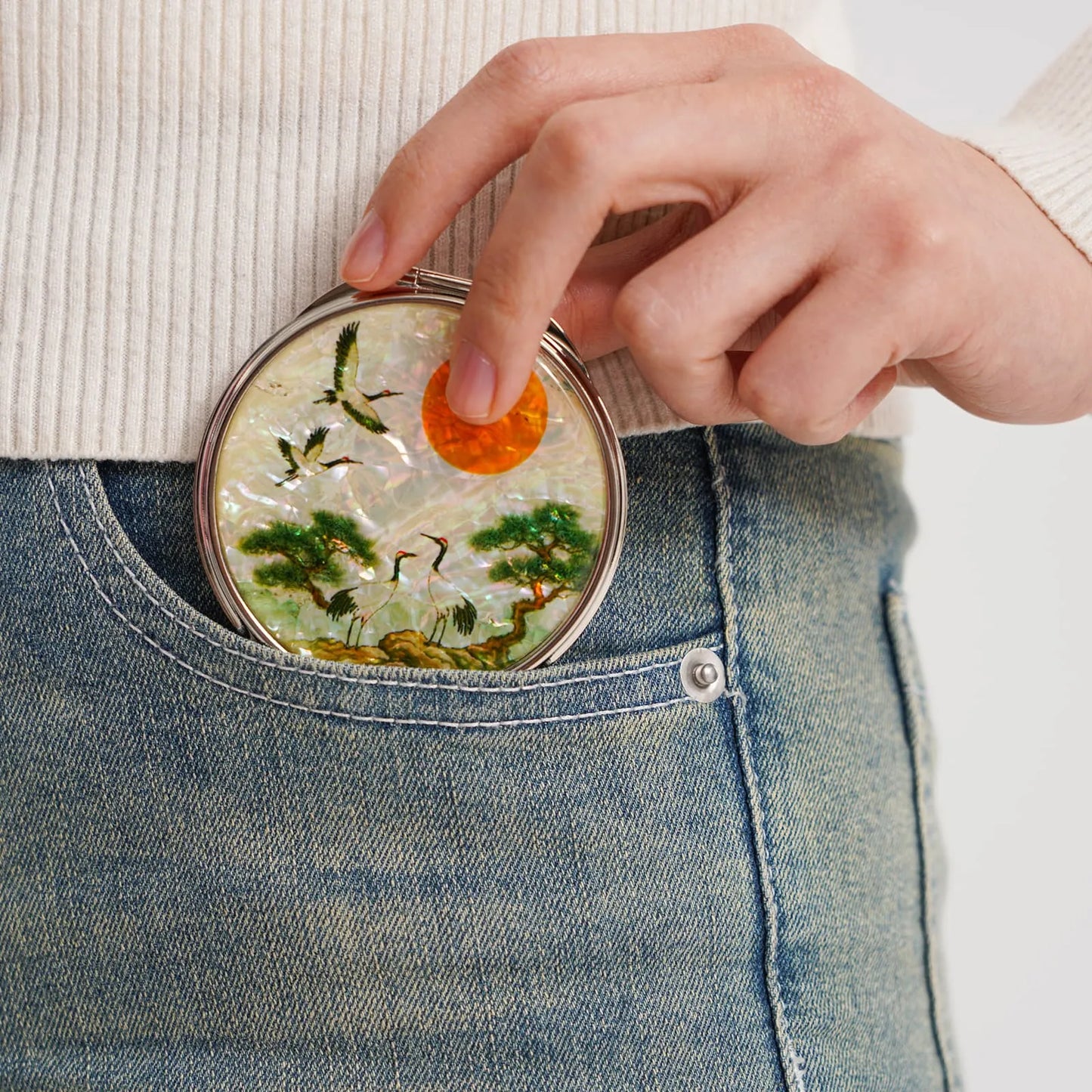 A hand placing a round compact mirror into the front pocket of blue jeans. The mirror features cranes, trees, and a bright orange sun in its design
