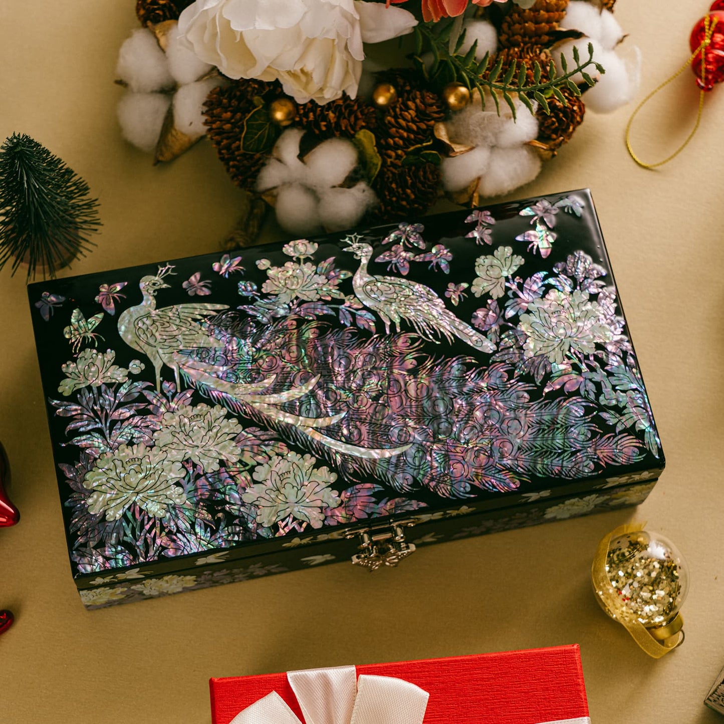 A jewelry box with a mother of pearl peacock design on a festive table, surrounded by holiday decorations, cotton branches, and a red gift box.