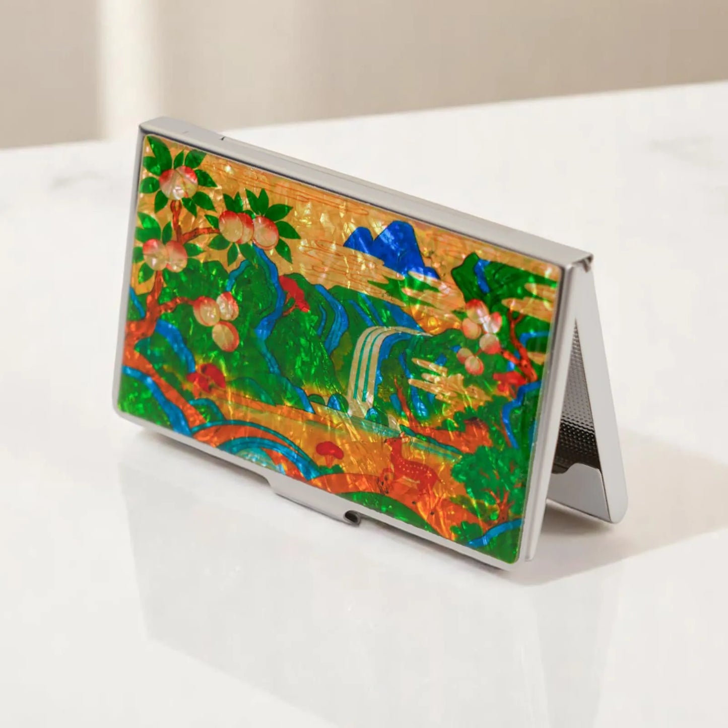 A metallic case featuring a colorful landscape with mountains, waterfalls, trees with fruit, and flowing rivers