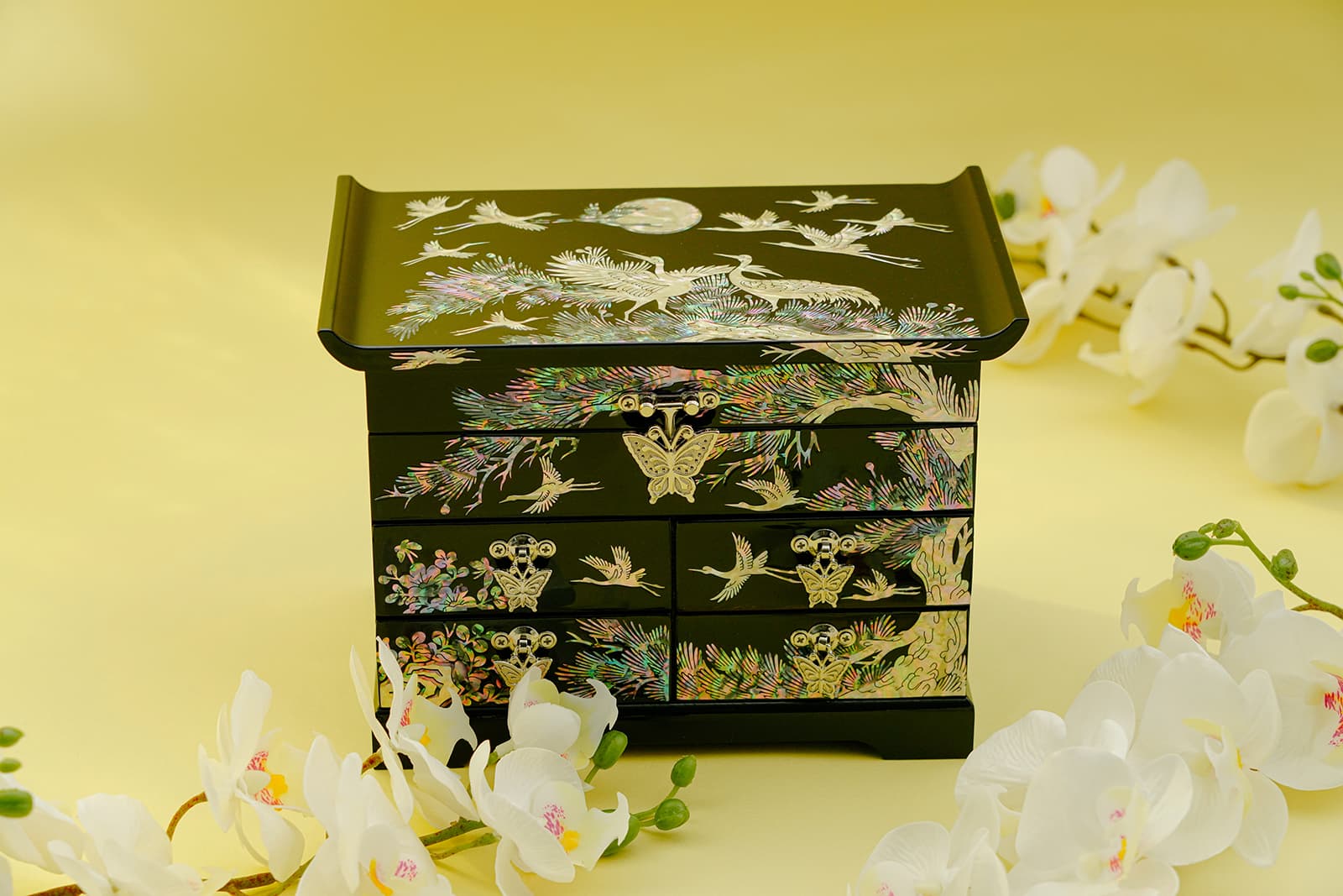 An exquisitely designed mother-of-pearl jewelry box featuring crane motifs, set against a yellow background with white orchids enhancing its beauty. The box's intricate details reflect traditional artistry, perfect for adding elegance to any space.