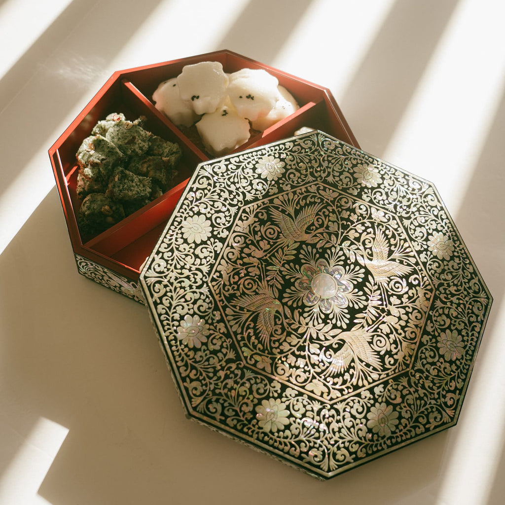 An octagonal Mother of Pearl inlaid box with a floral design, lid closed, next to its open base filled with Korean sweets, in sunlight