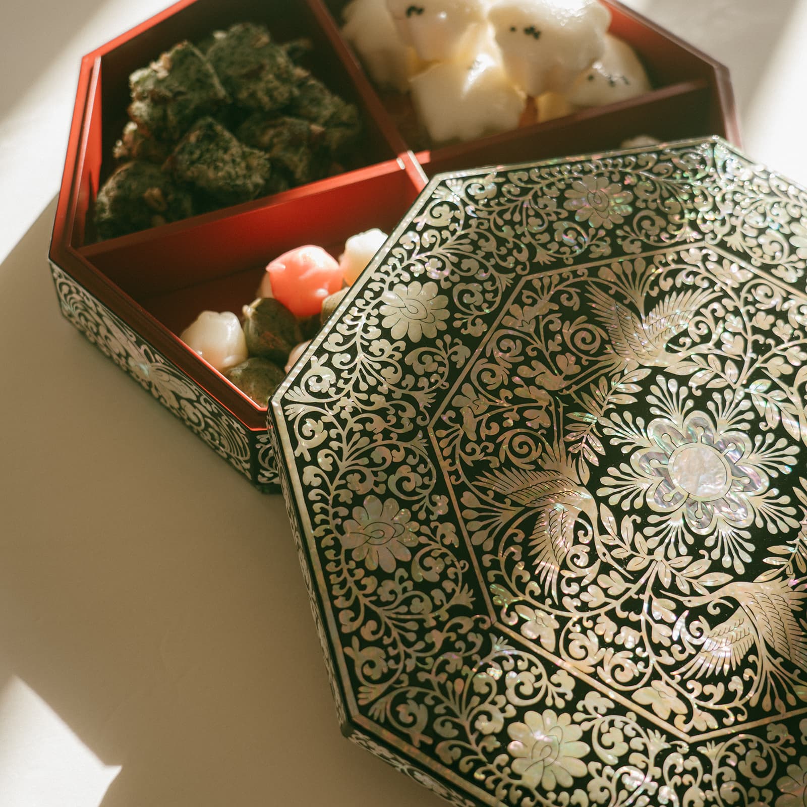 An octagonal Mother of Pearl inlaid box with the lid placed beside the base, which is filled with colorful Korean confections, in a sunlit setting