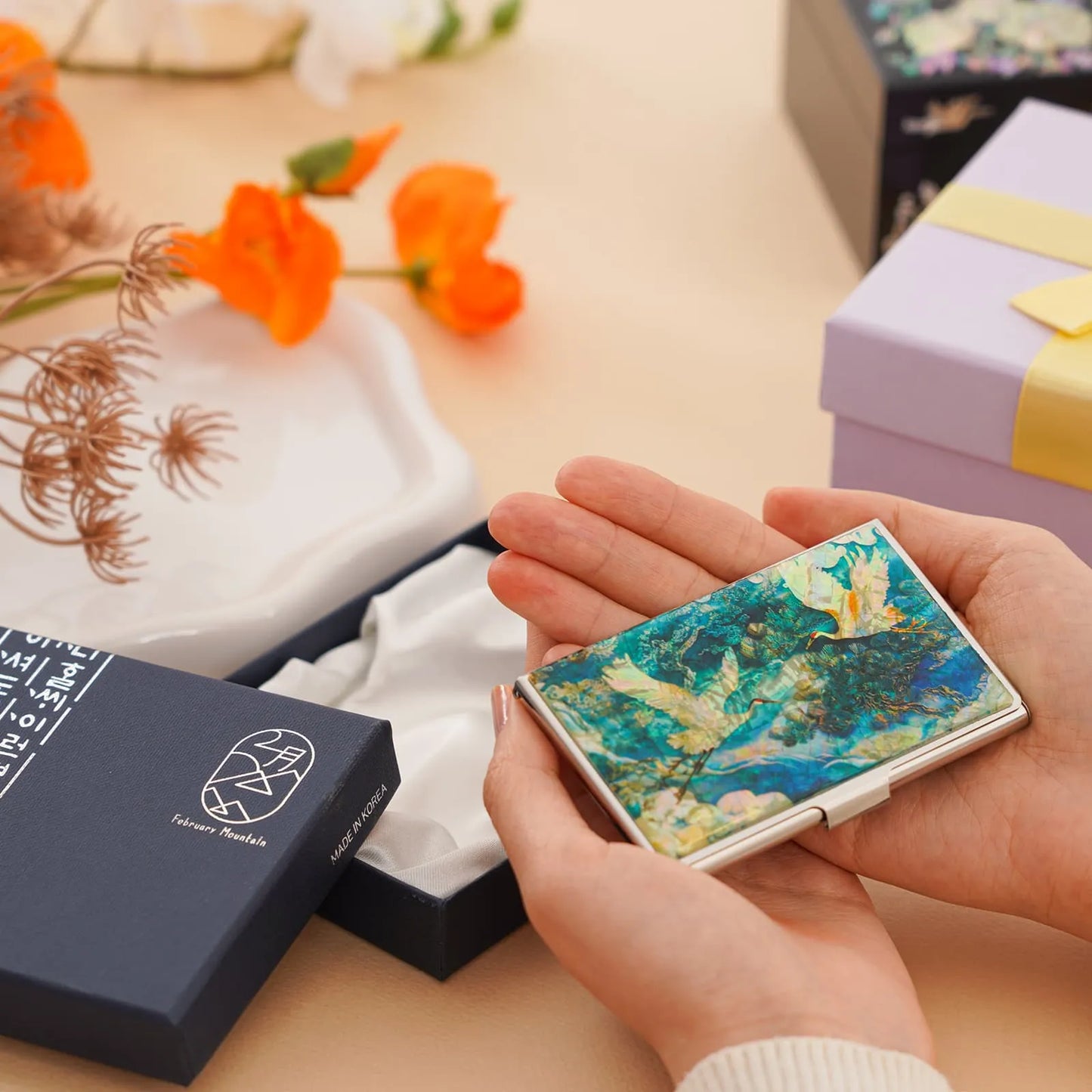 Person holding a decorative cardholder with floral design, surrounded by gift boxes and flowers on a table.