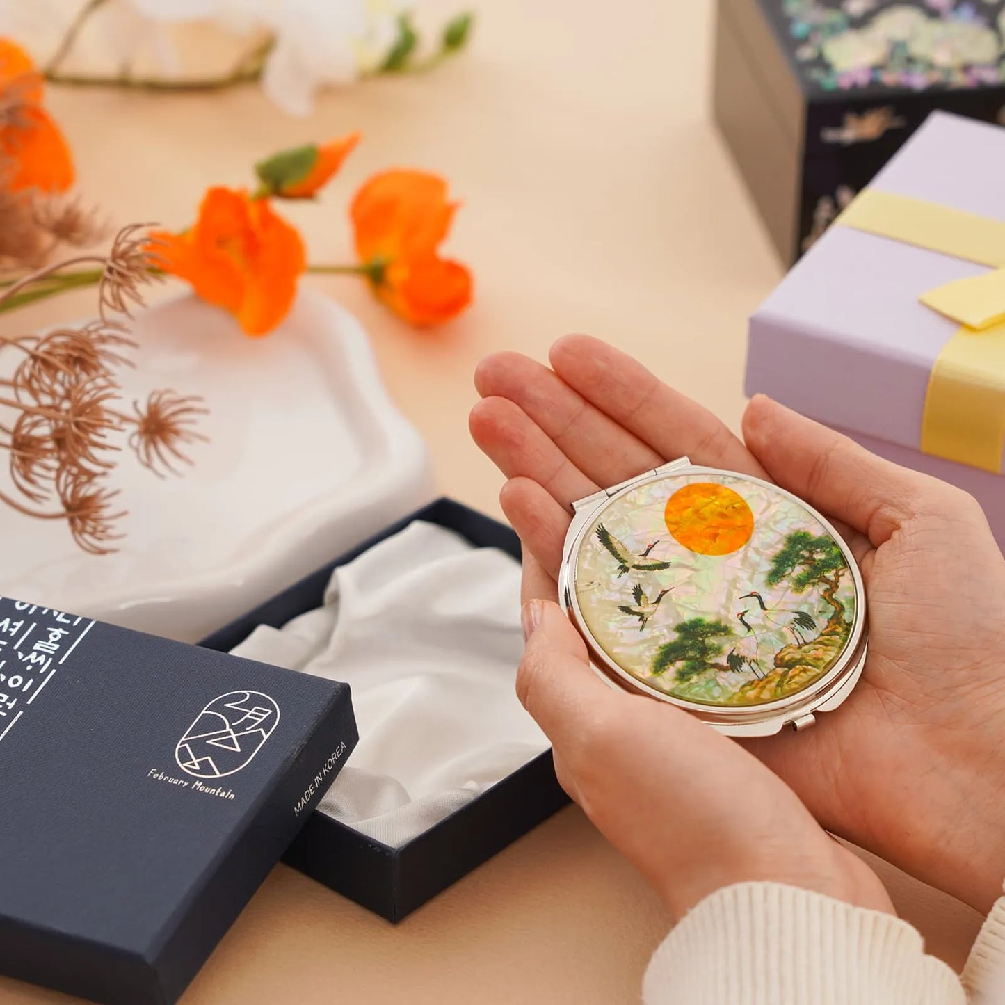 A person holds a decorative compact mirror with a nature scene, surrounded by gift boxes and flowers