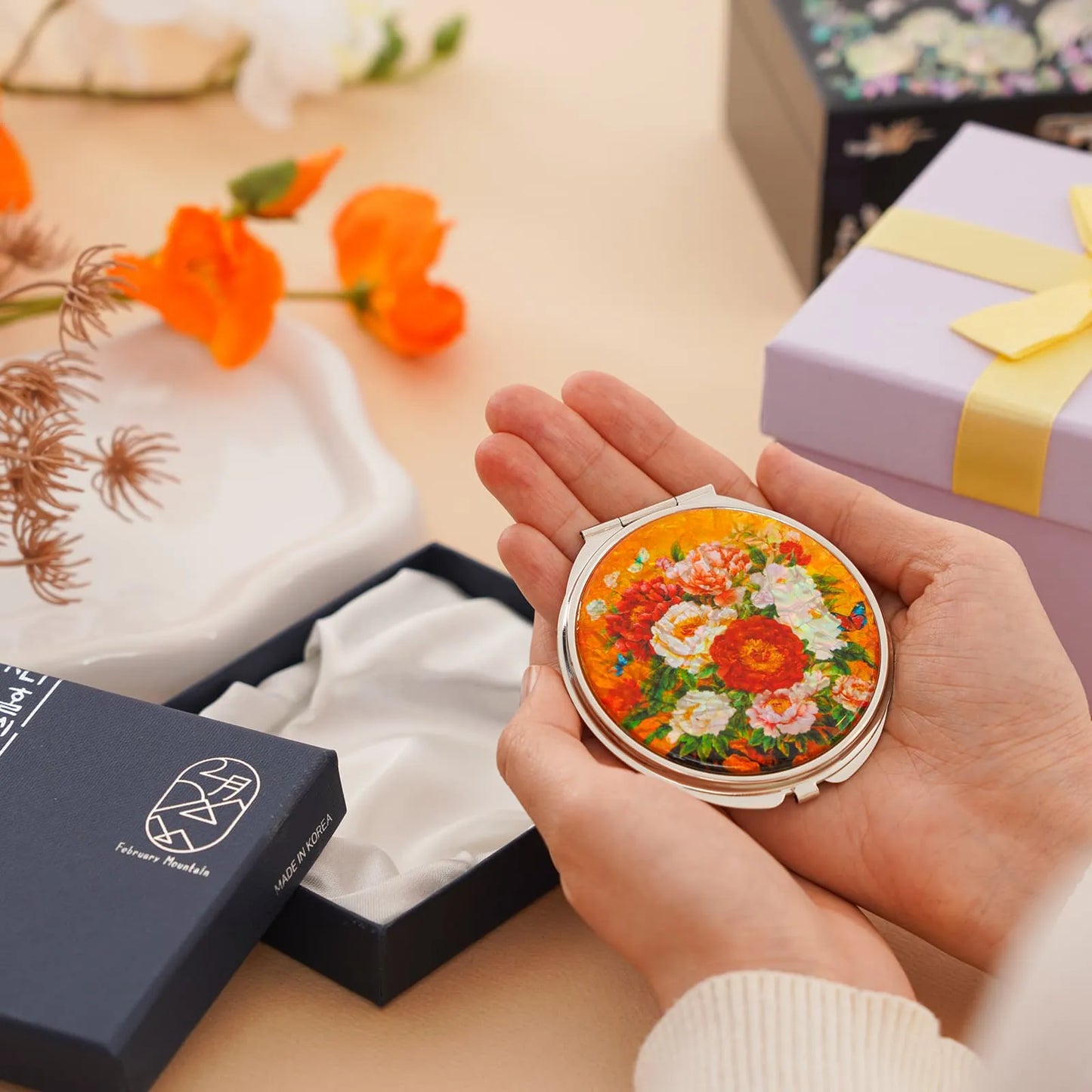A person holds a round compact mirror with a vibrant floral design featuring red, white, and pink flowers against an orange background