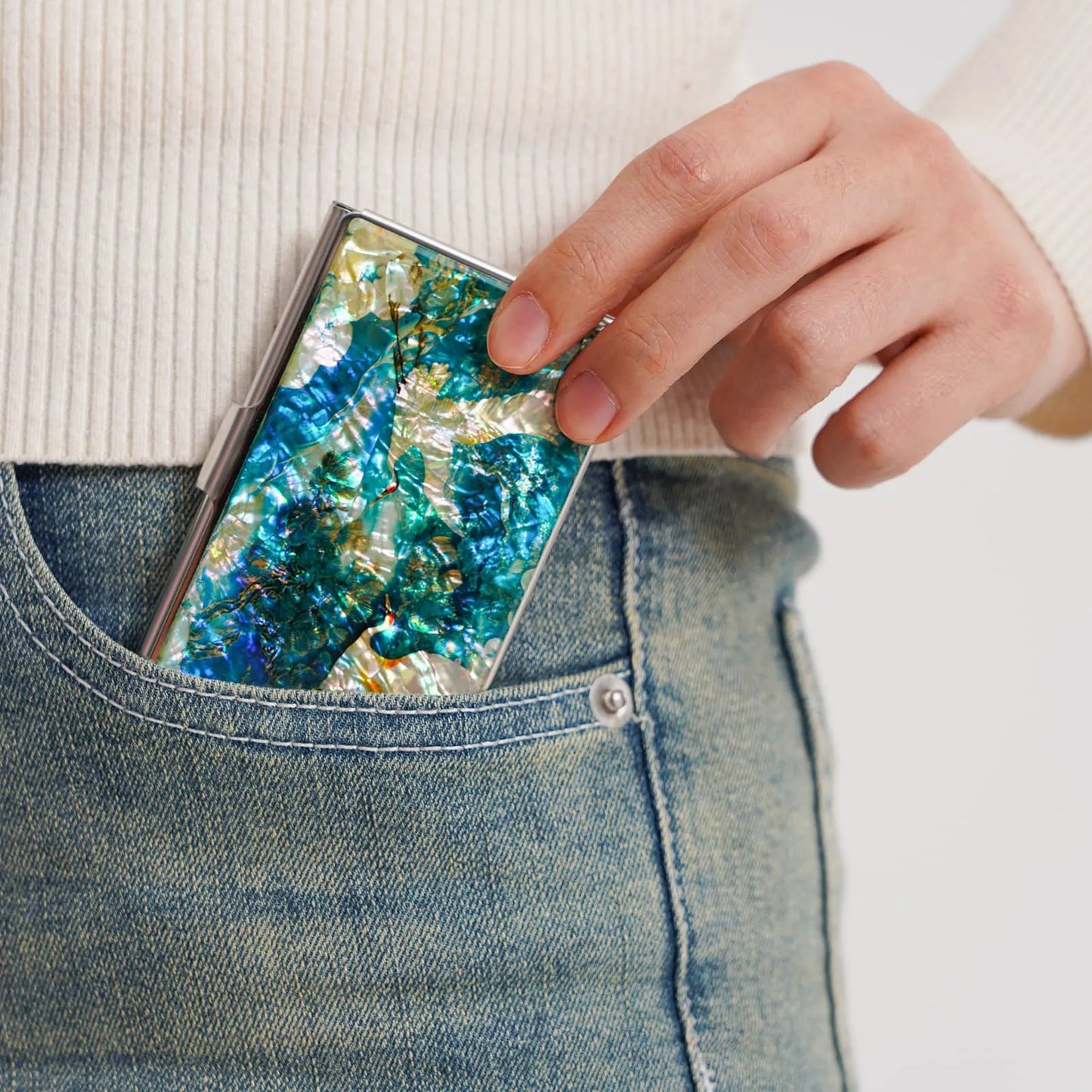 A person places a colorful, iridescent case with blue, green, and gold hues into a blue jean pocket