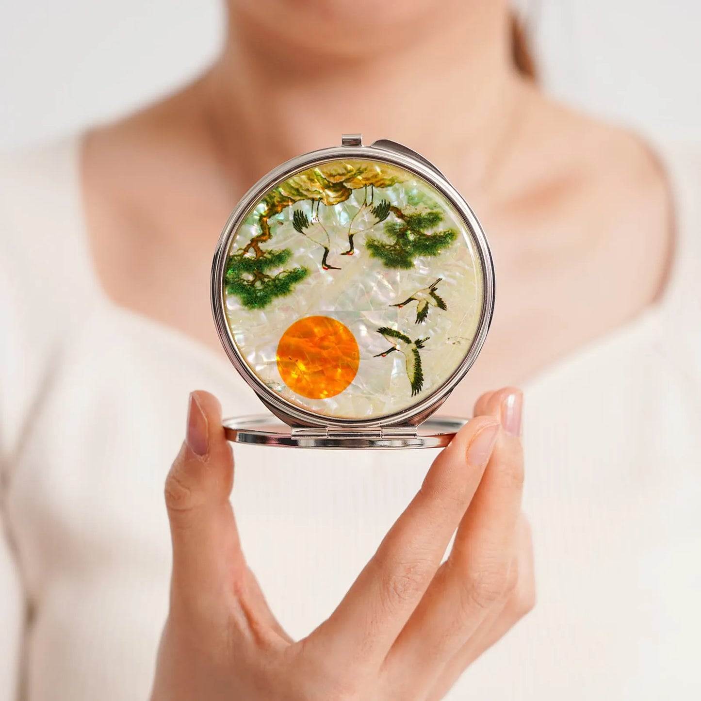 A round compact mirror featuring an intricate design with cranes, pine trees, and a glowing orange sun