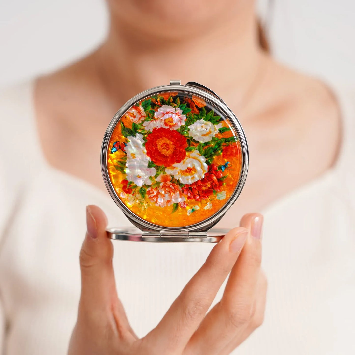 A round compact mirror featuring a vibrant floral design with orange, red, and white flowers against a bright orange background, held by a person wearin