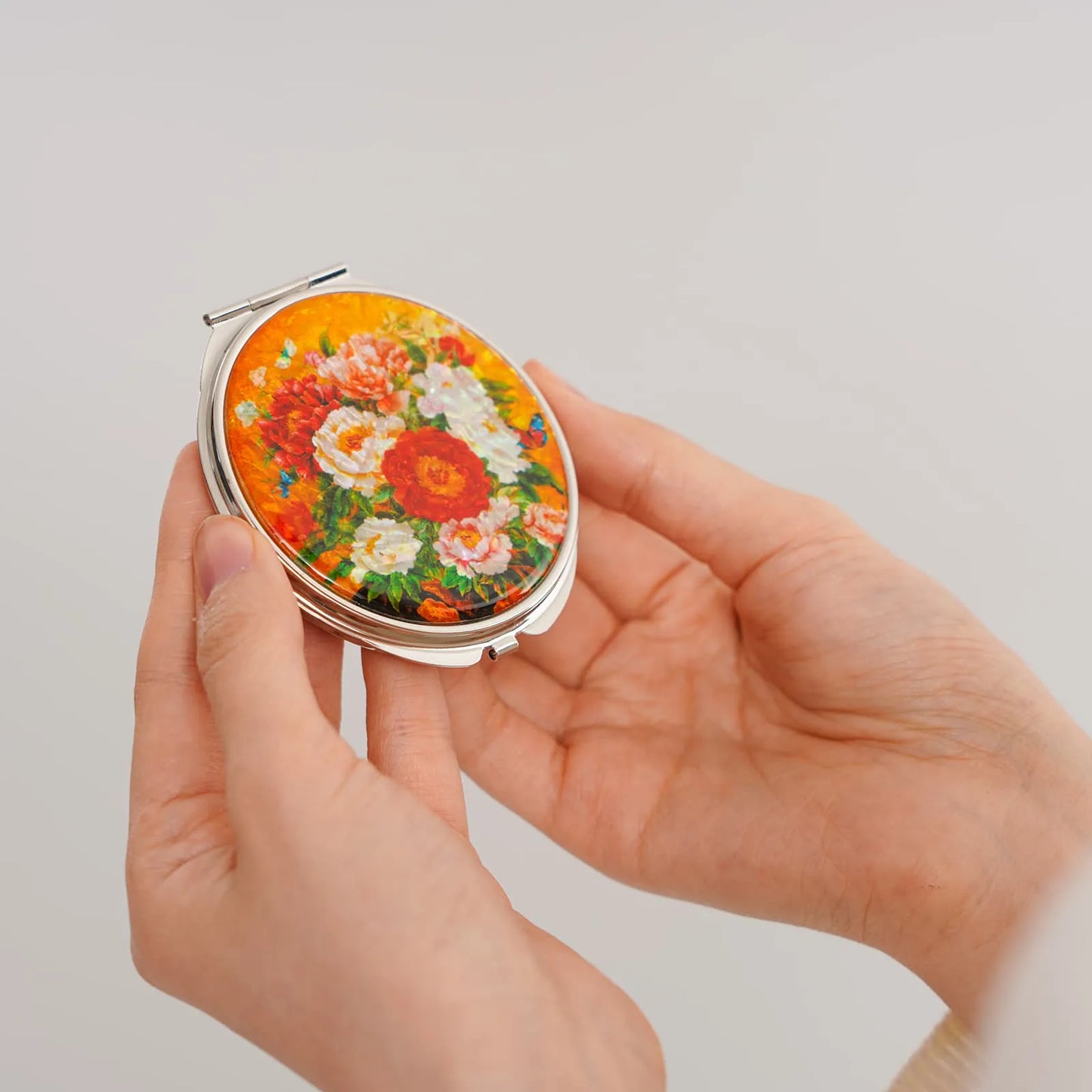 A round compact mirror featuring a vibrant floral design with red, white, and pink flowers against a yellow background, held in someone’s hands