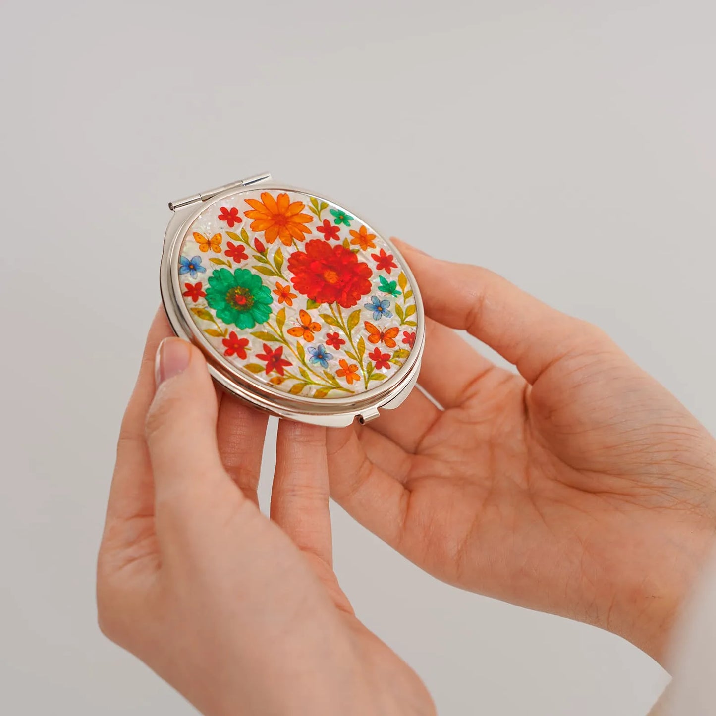 A round compact mirror featuring a vibrant floral pattern, held above an open satin-lined box