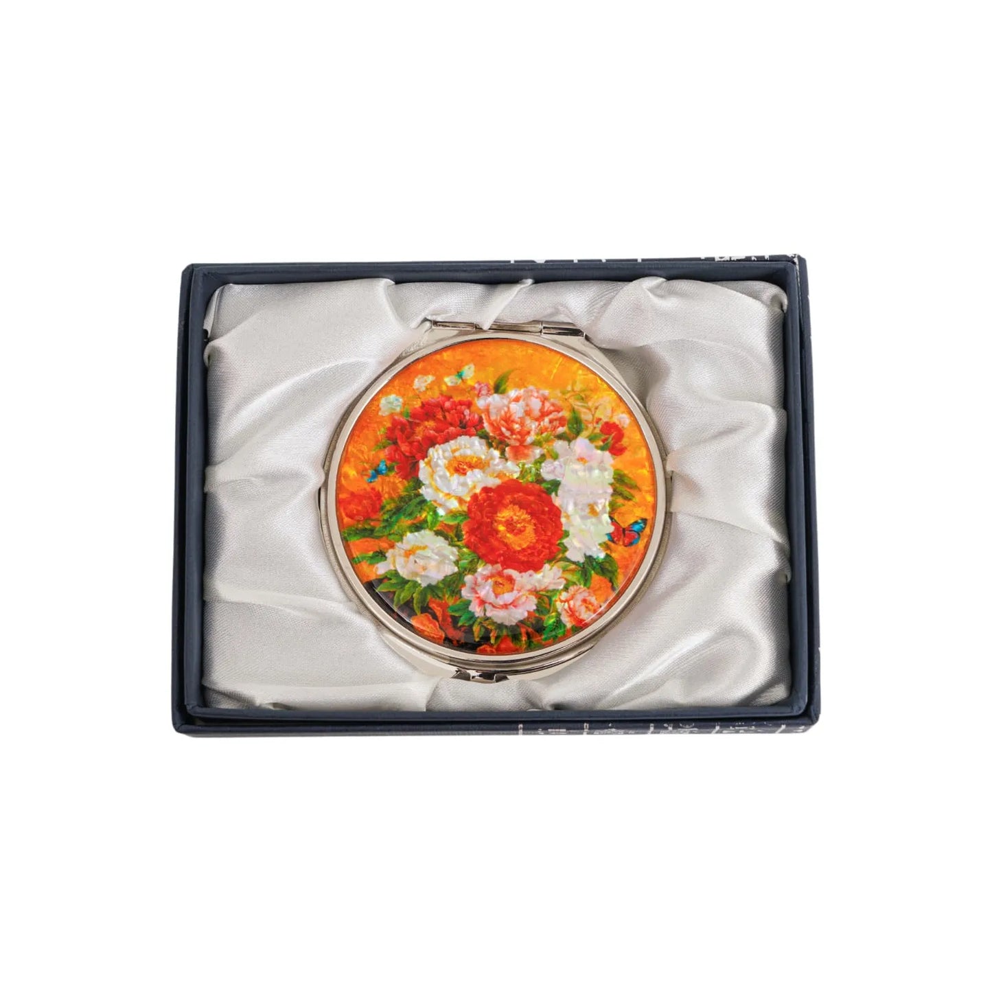 A round compact mirror with a colorful floral design featuring red, white, and orange flowers against a yellow background, placed inside a black satin-l