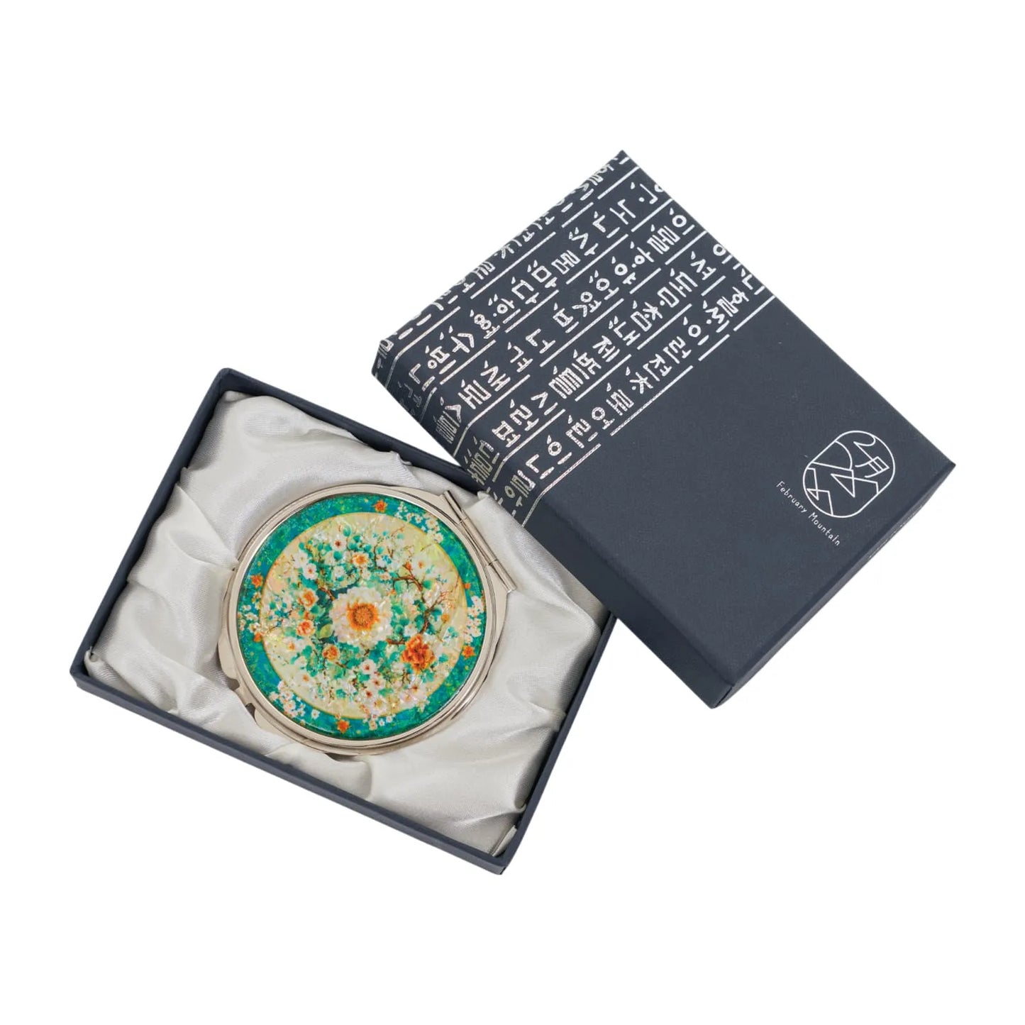 A round compact mirror with a decorative floral design featuring white, orange, and yellow flowers against a green and yellow background. The mirror is (