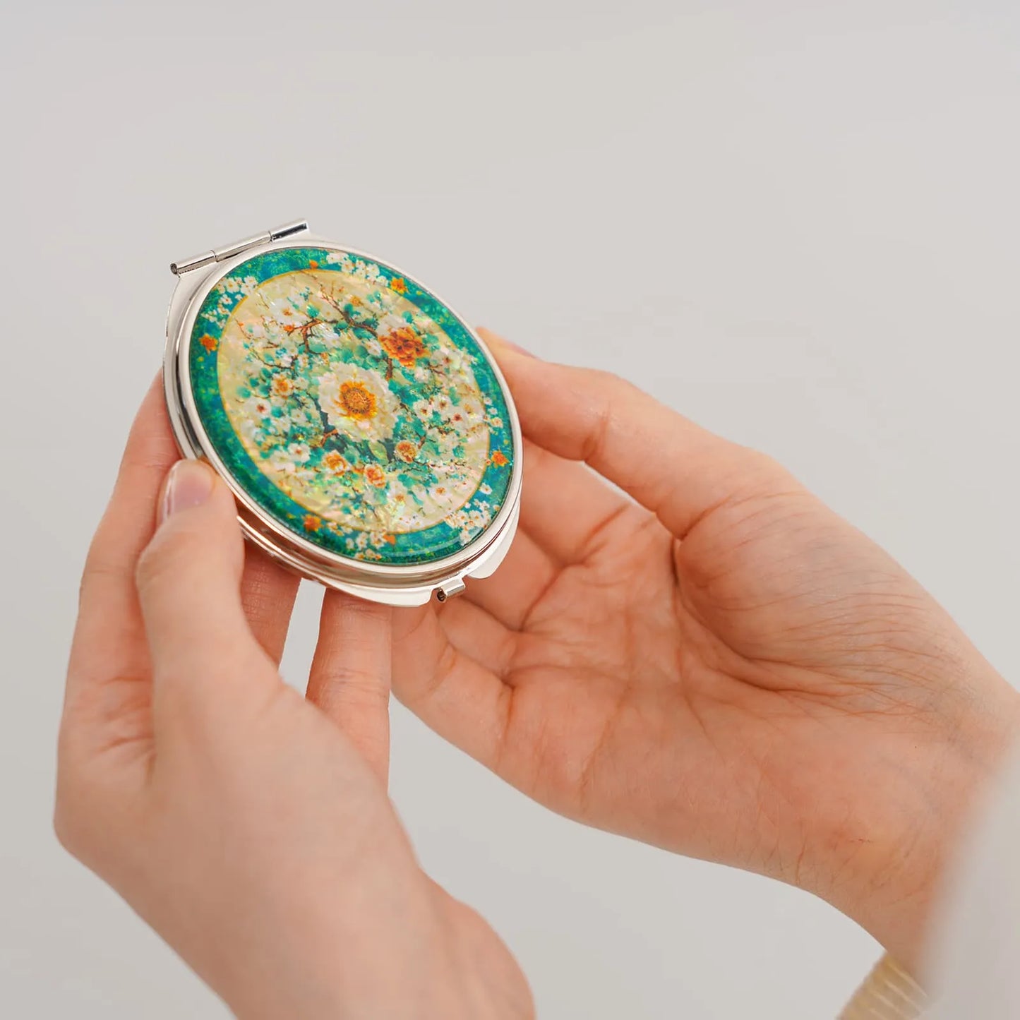 A round compact mirror with a decorative floral design featuring white, orange, and yellow flowers against a green and yellow background. The mirror is ( (3)