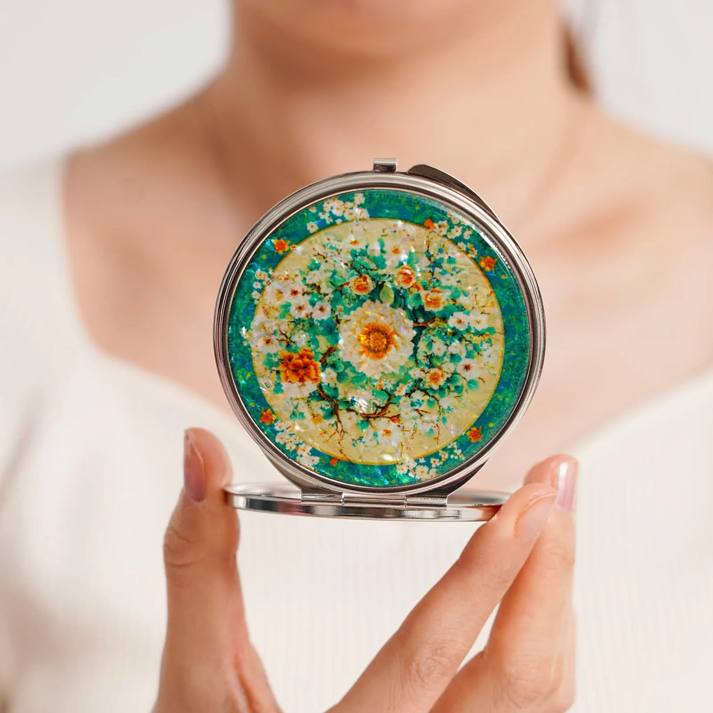 A round compact mirror with a decorative floral design featuring white, orange, and yellow flowers against a green and yellow background. The mirror is ( (5)