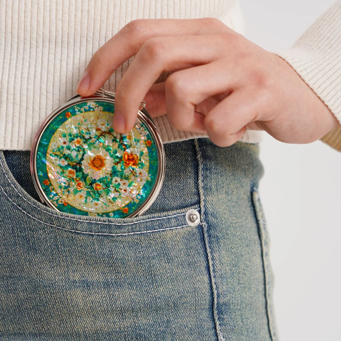 A round compact mirror with a decorative floral design featuring white, orange, and yellow flowers against a green and yellow background. The mirror is ( (6)