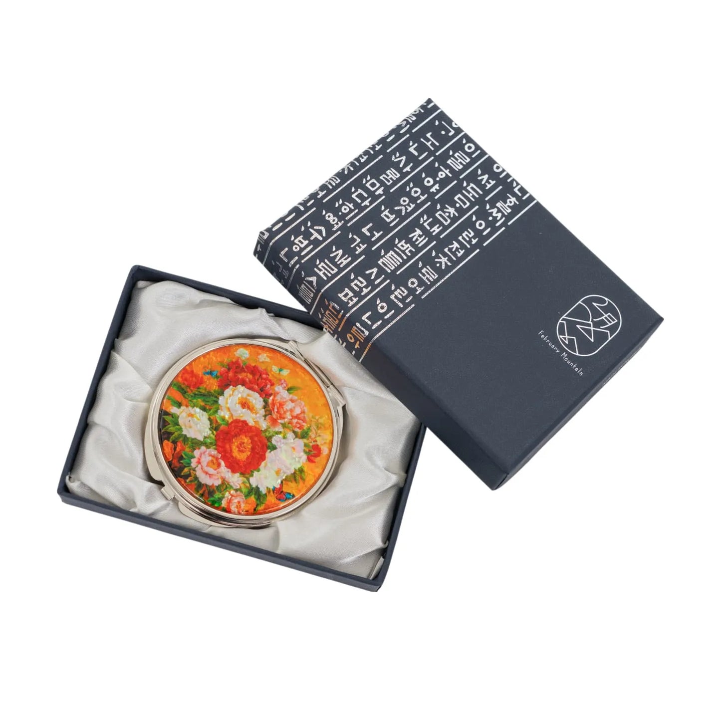 A round compact mirror with a floral design featuring red, pink, and white flowers on an orange background, placed inside a satin-lined dark blue box wi
