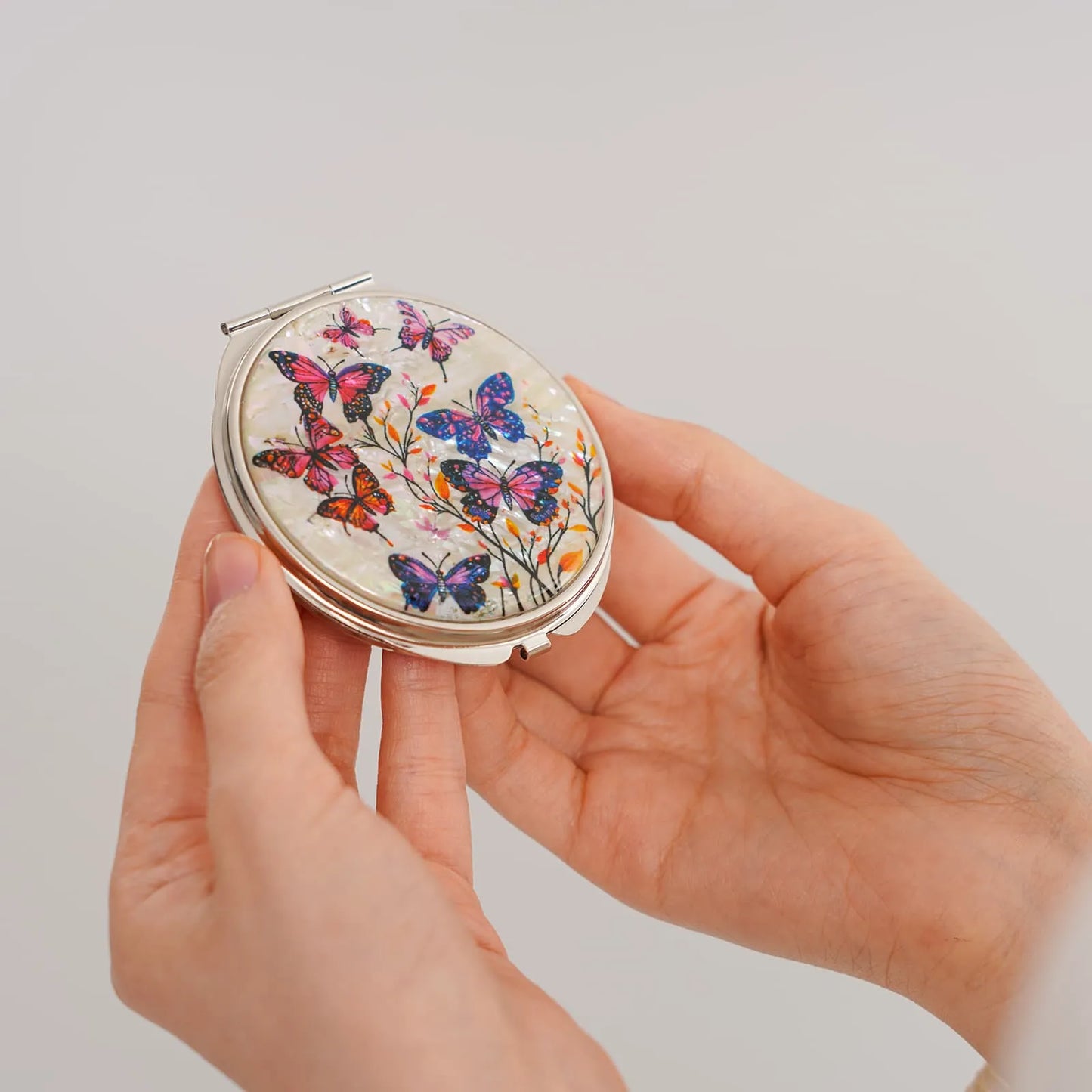 A round compact mirror with an intricate butterfly and floral design in shades of pink, purple, blue, and orange, held in someone's hand