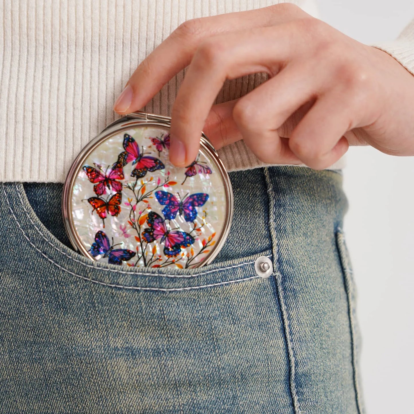 A round compact mirror with a vibrant butterfly design in shades of red, pink, purple, and blue, being placed into the front pocket of blue jeans