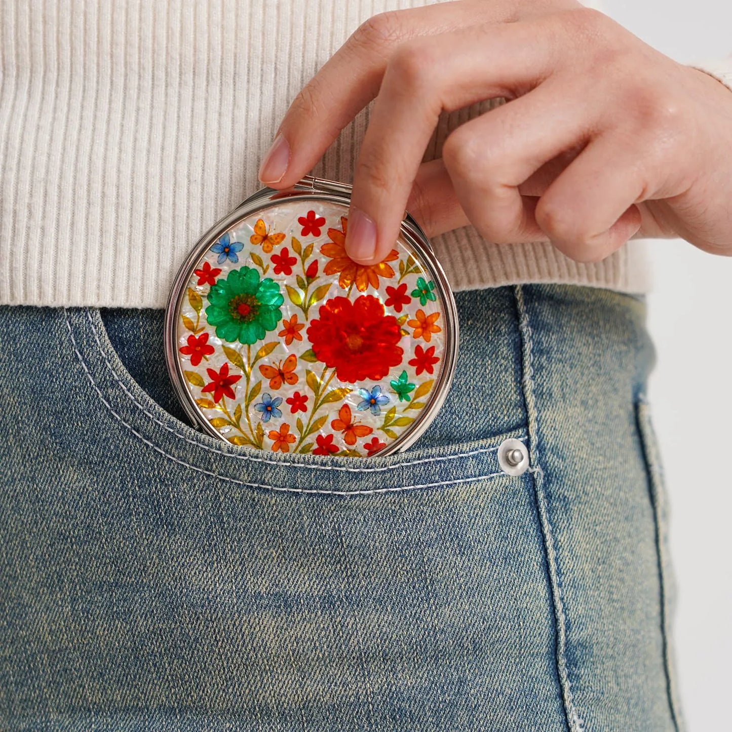 A round compact mirror with a vibrant floral design, being placed into the front pocket of blue jeans