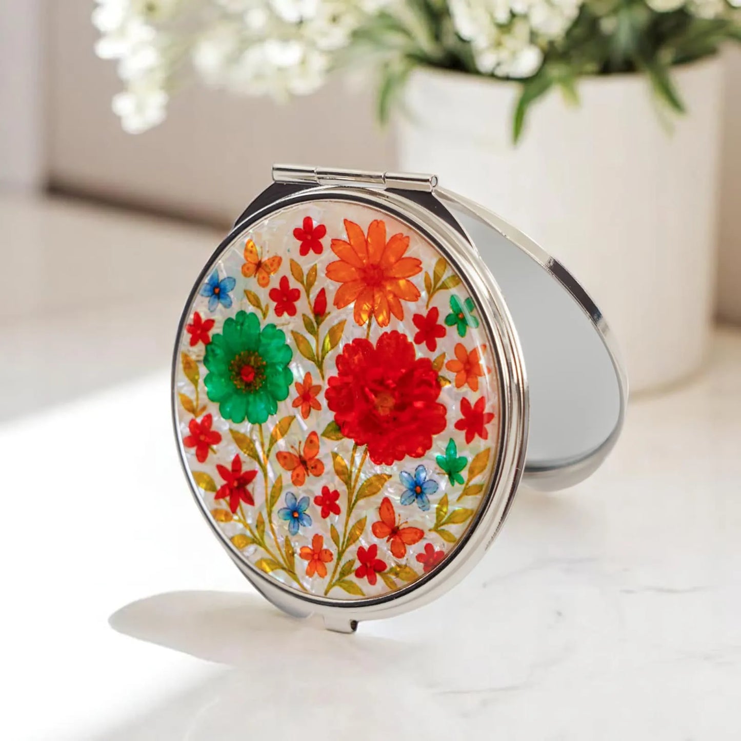 A round compact mirror with colorful floral designs, including red, green, and orange flowers, on its cover.