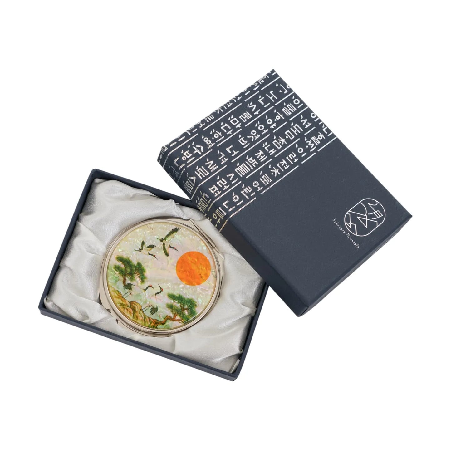 A round decorative item with an intricate design of cranes, trees, and a glowing orange sun, placed inside a dark blue box lined with white satin fabric.