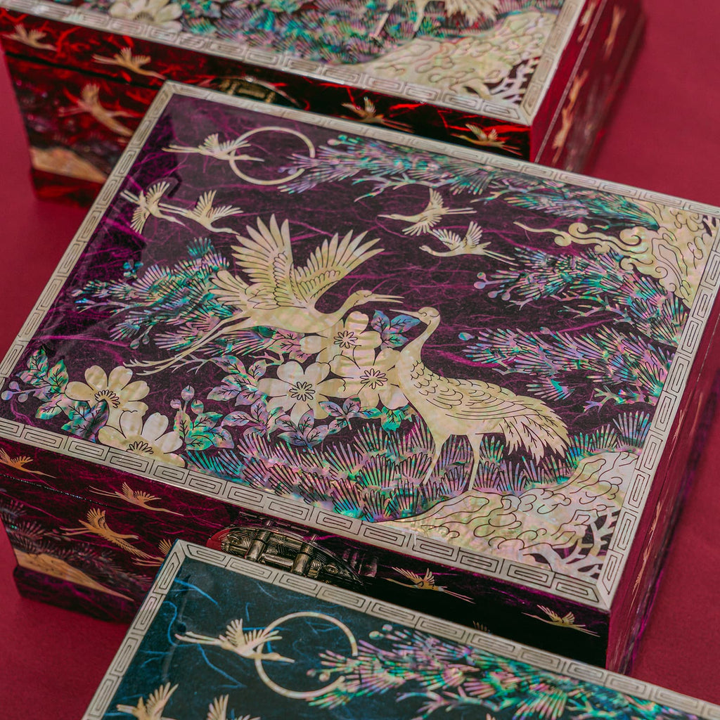 A set of Mother of Pearl boxes with detailed crane and floral designs on a burgundy background, displayed on a table.