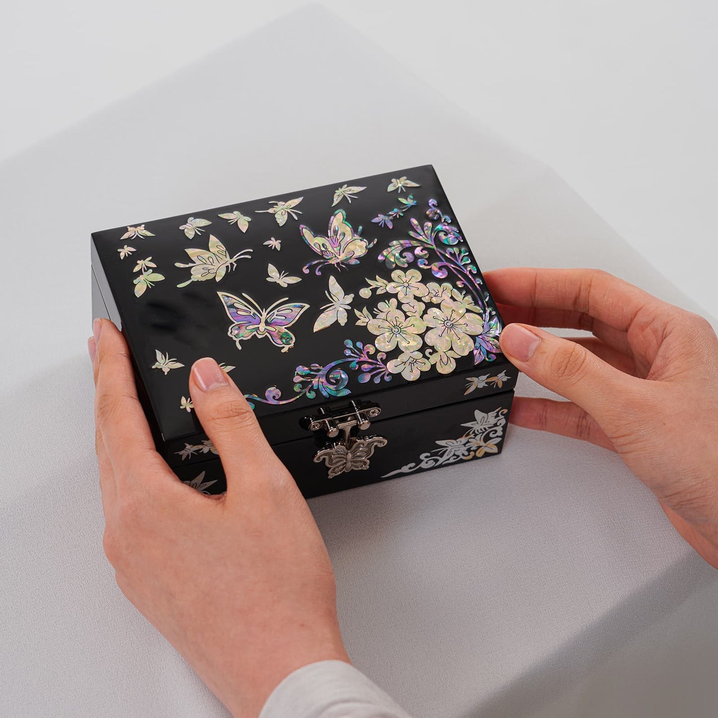 Black jewelry box with colorful butterfly inlay, held in two hands over light fabric surface