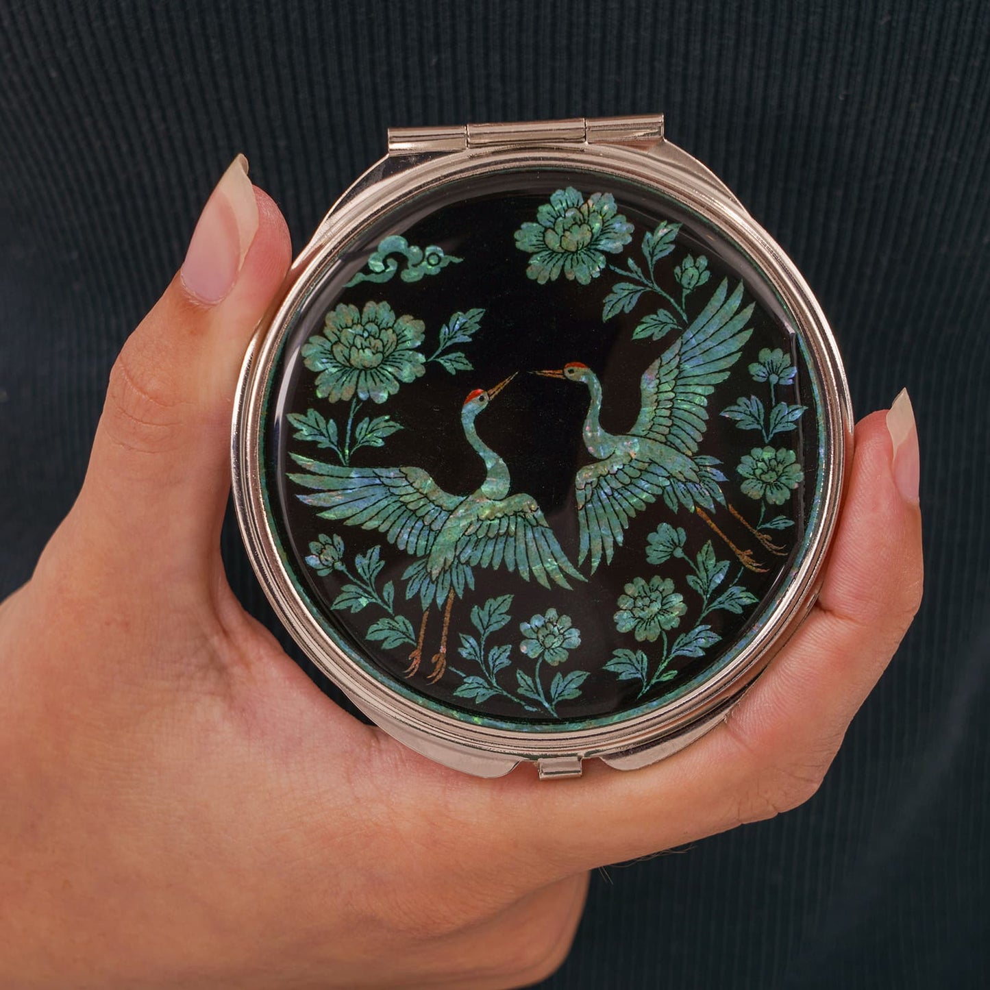 Black compact mirror with twin cranes and peonies in hand over dark background