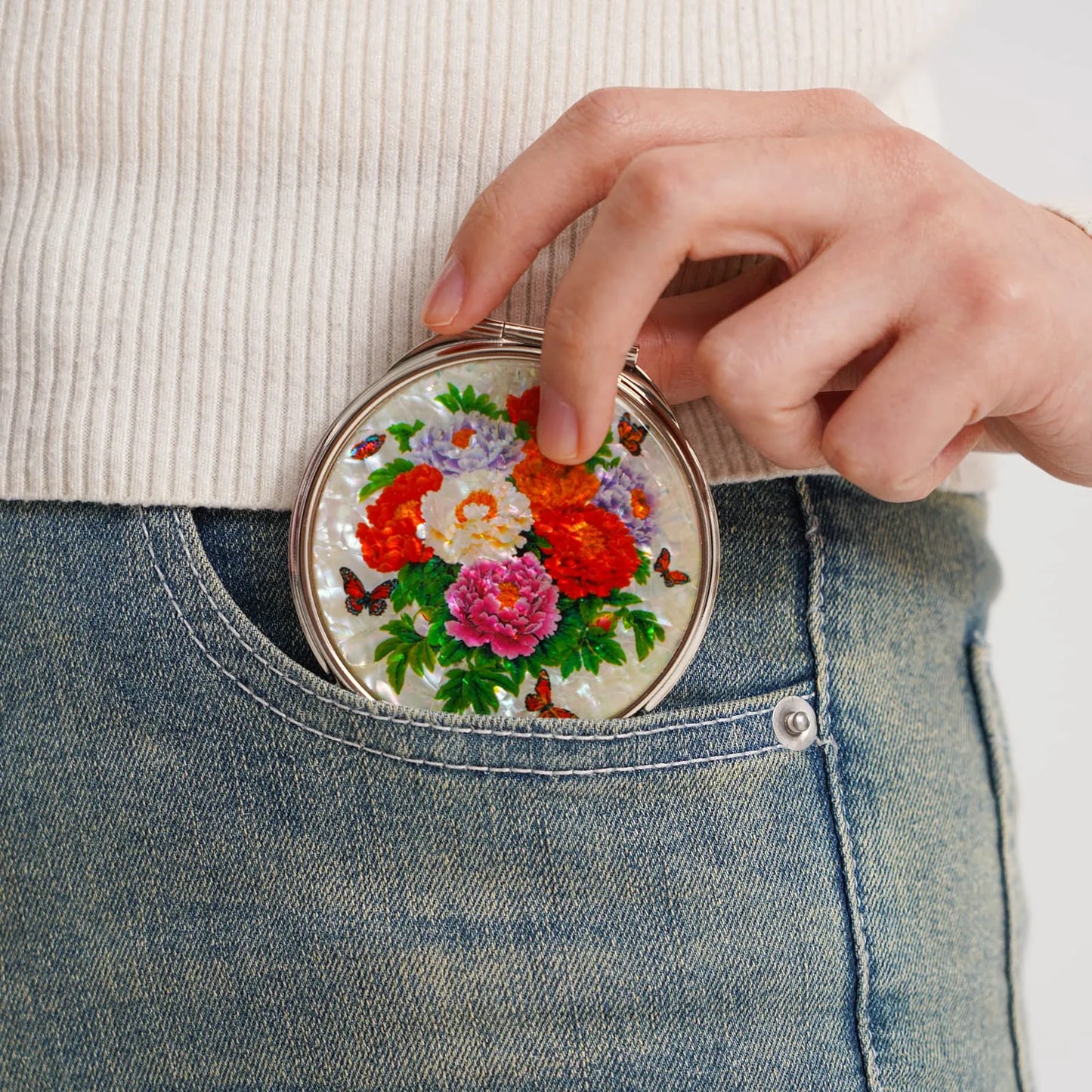Compact mirror featuring colorful peonies being placed into a jeans pocket