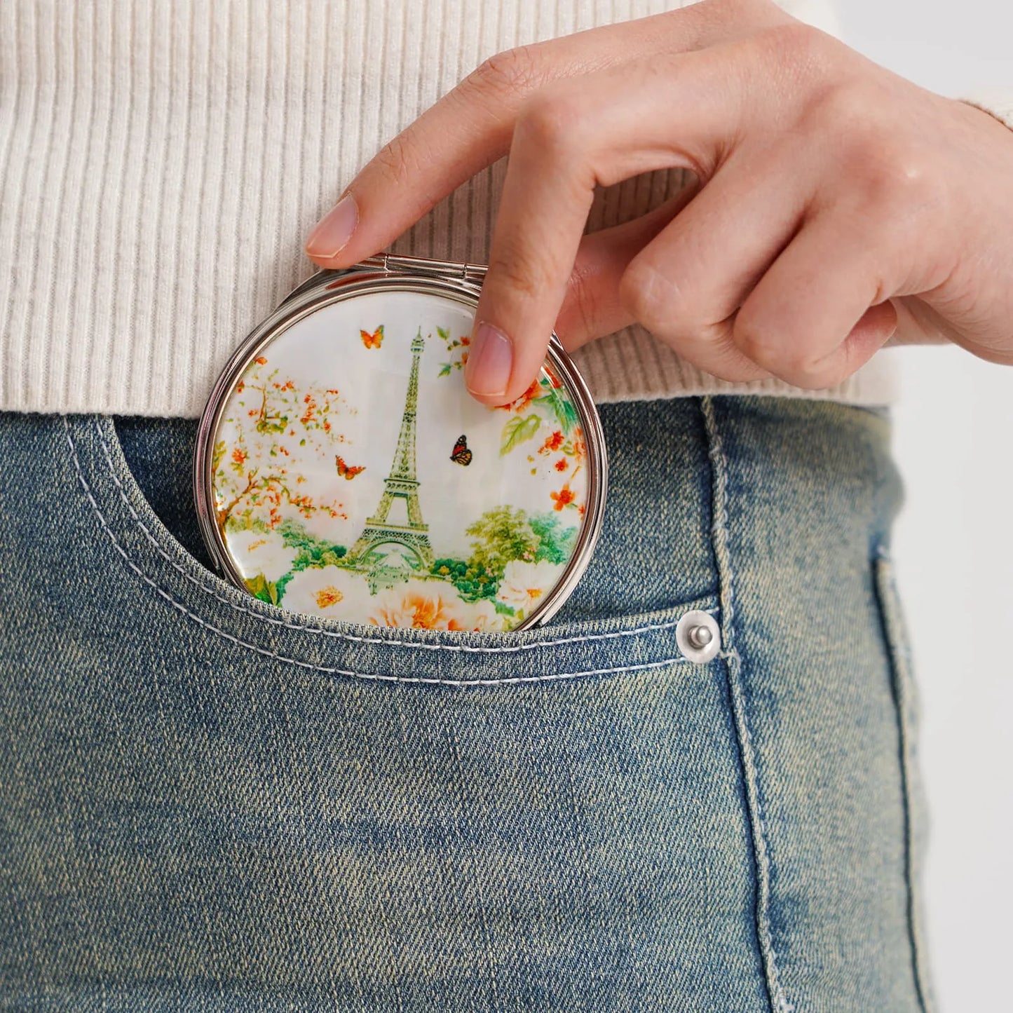 Compact mirror with Eiffel Tower, flowers, and butterflies being placed into the front pocket of blue jeans