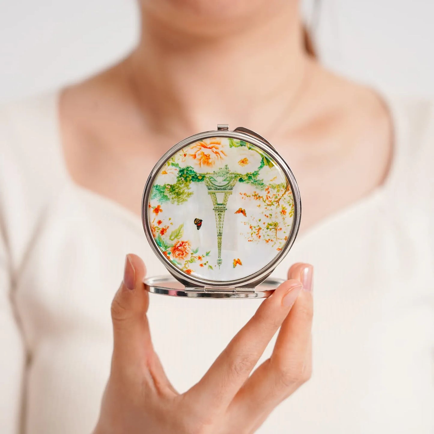 Compact mirror with elegant green and orange floral design, held by a person wearing a white top