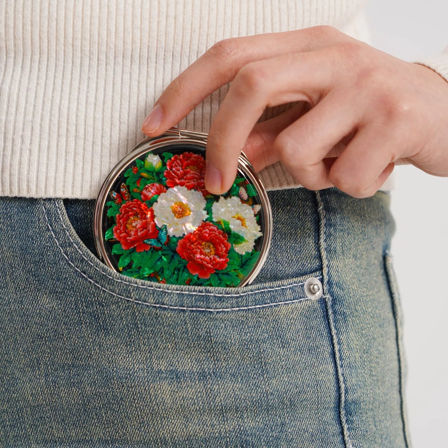 Compact mirror with peonies being placed into a jeans pocket