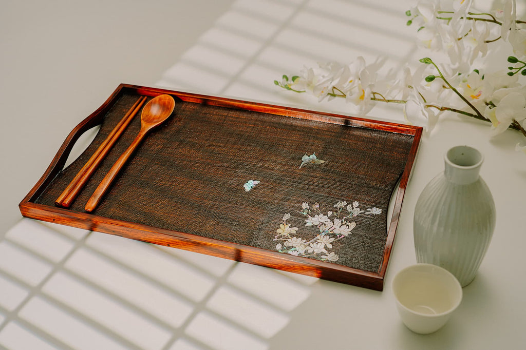 Elegant rectangular tray with dark, textured surface, adorned with delicate mother-of-pearl inlay of butterflies and flowers in one corner. Traditional wooden utensils rest on top, with a serene
