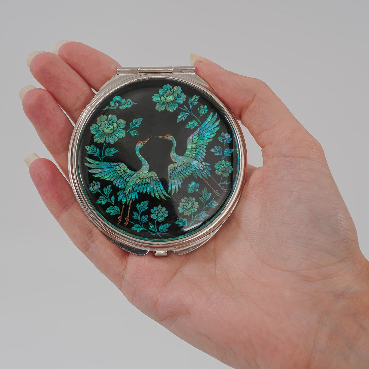 Hand holding compact mirror with cranes and peony floral design on black mother-of-pearl finish