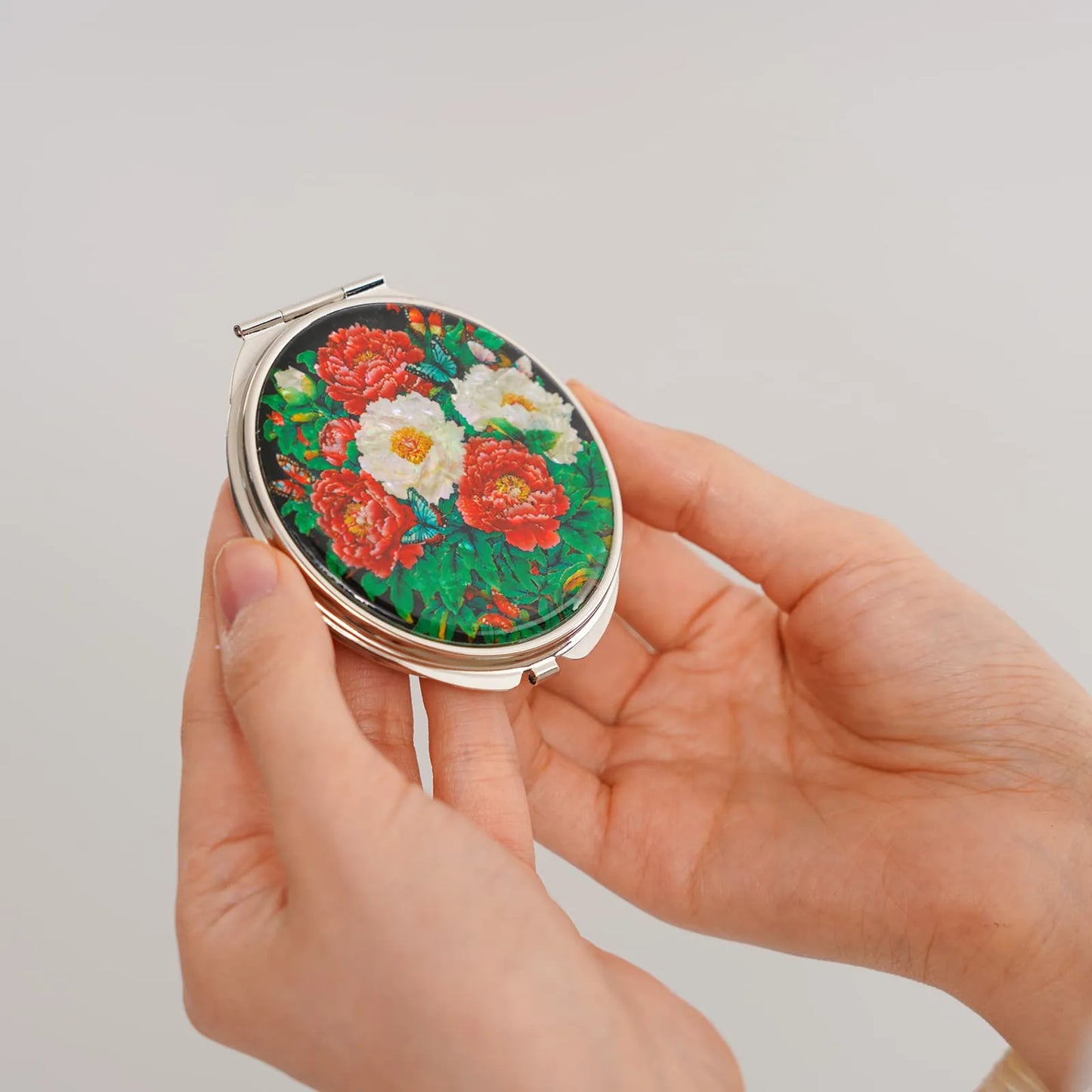 Hands holding a compact mirror with mother-of-pearl floral inlay on a plain background
