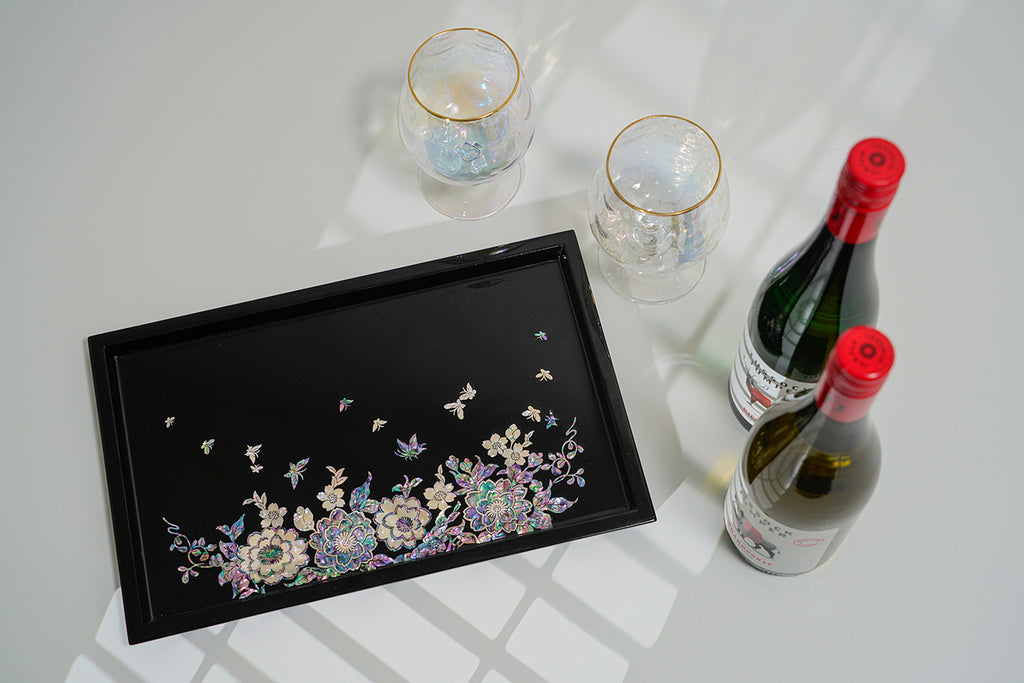 This image shows a glossy black mother-of-pearl tray with a detailed inlay of flowers and butterflies, accompanied by two iridescent wine glasses and two bottles of wine, placed on a white surface with light casting geometric shadows.