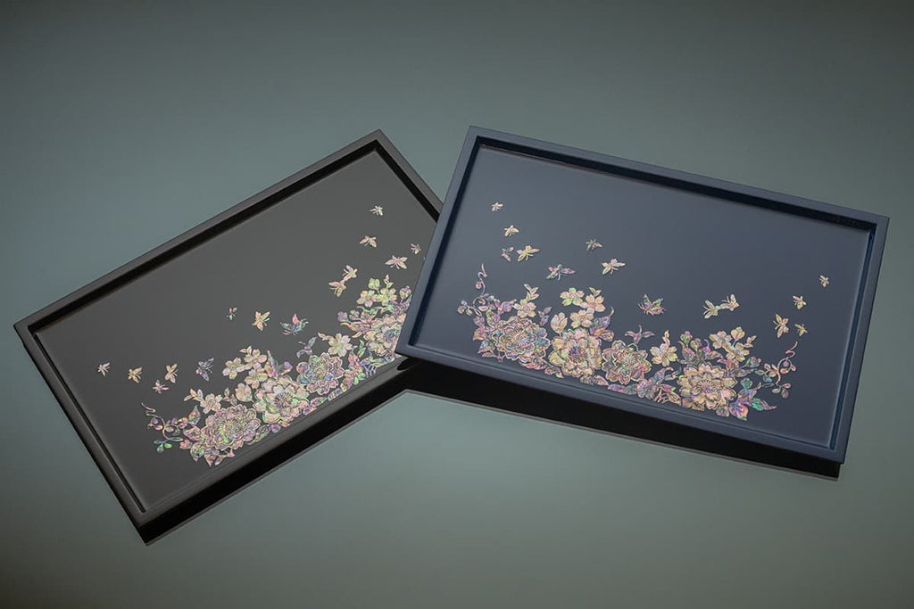 Two rectangular trays with dark backgrounds, each featuring a colorful Mother of Pearl inlay of floral and butterfly designs, overlapping and presented against a muted backdrop.