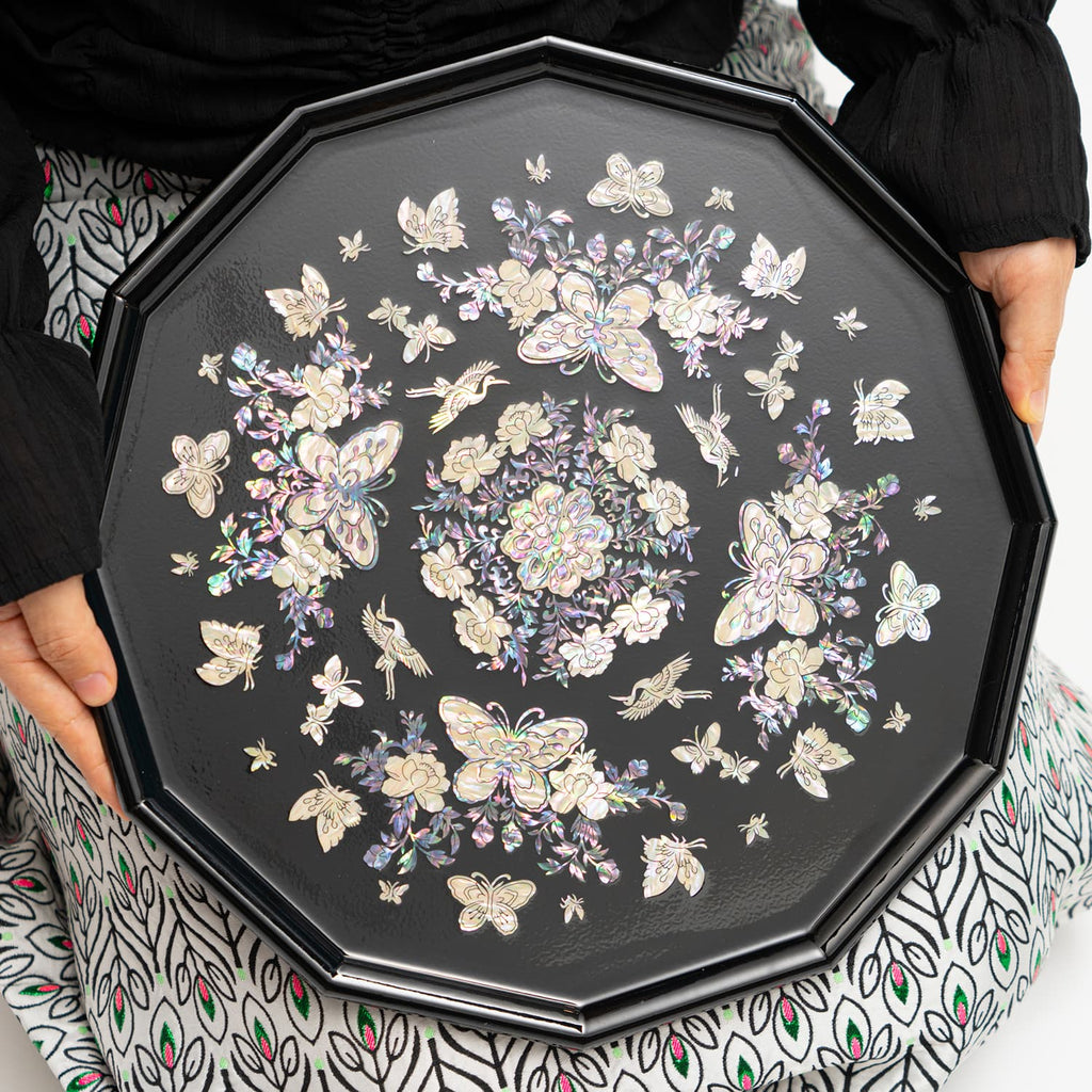 The image showcases an octagonal mother-of-pearl tray with intricate floral and butterfly motifs, being held by someone.
