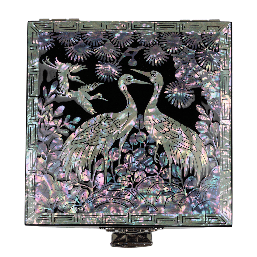 An exquisite jewelry box featuring mother-of-pearl cranes and pine tree design on a black backdrop, highlighted by a patterned frame, mounted on a pedestal.