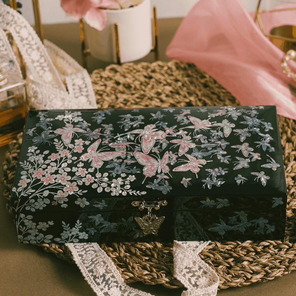 A black lacquered jewelry box with a mother of pearl floral and butterfly motif, set on a woven table mat with delicate lace and romantic decor in the background.
