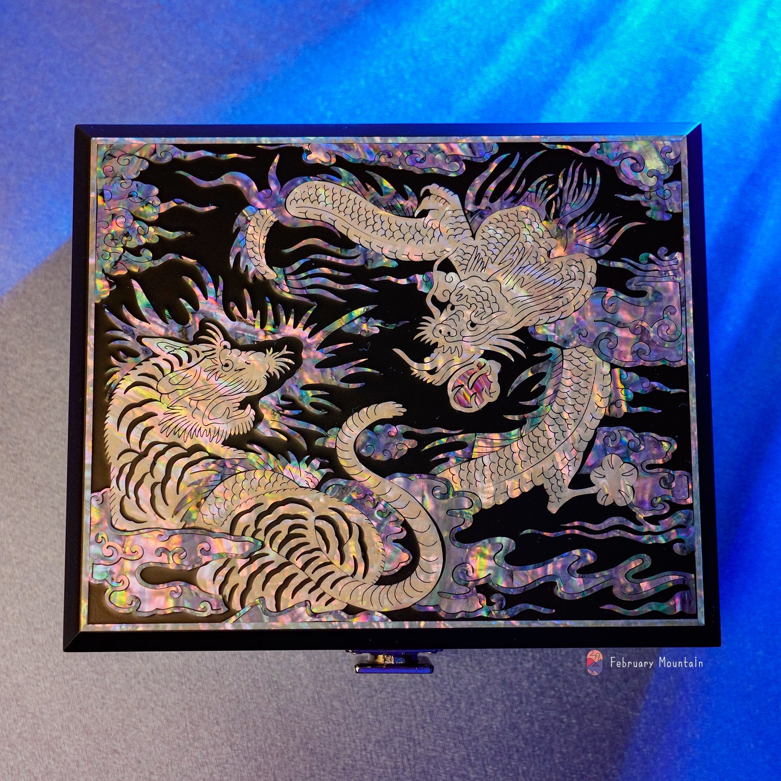 A decorative box with a tiger and dragon mother of pearl inlay, displayed against a blue light, with the February Mountain logo.