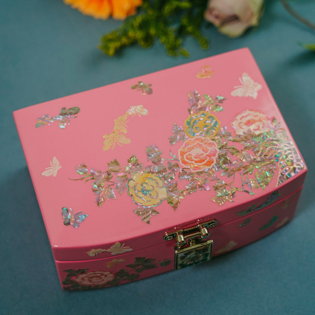 A soft pink jewelry box is showcased against a muted blue backdrop. The box is embellished with delicate floral motifs, butterflies, and shimmering accents that catch the light. In the blurred background, an arrangement of vibrant flowers – an orange gerbera daisy, a purple rose, and a pale pink rose – along with some greenery, complements the main subject. The gentle pastel tones of the scene create a dreamy and elegant atmosphere.