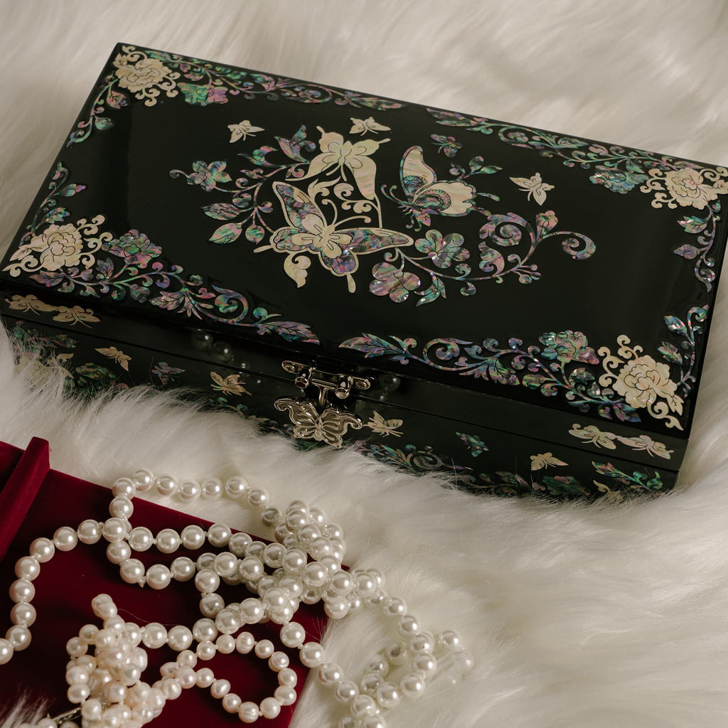 A black jewelry box adorned with mother-of-pearl inlays, showcasing a symphony of butterflies and florals in iridescent colors, is elegantly displayed against a soft, white furry backdrop, complemented by a string of pearls laid out in front.